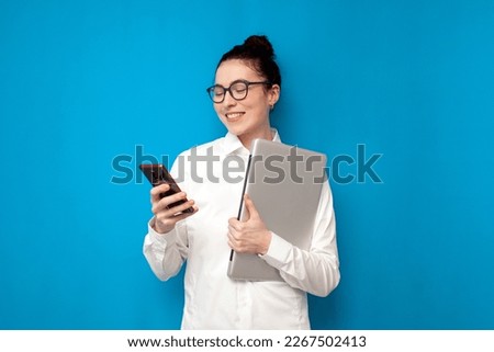 successful business woman in white shirt and glasses holds laptop and uses smartphone on blue background, girl manager financier is typing on phone on colored background, portrait of office worker Royalty-Free Stock Photo #2267502413