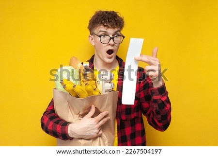 young shocked shopper with a package of groceries holding a check and being surprised by the high prices on a yellow background, the concept of a rise in price Royalty-Free Stock Photo #2267504197