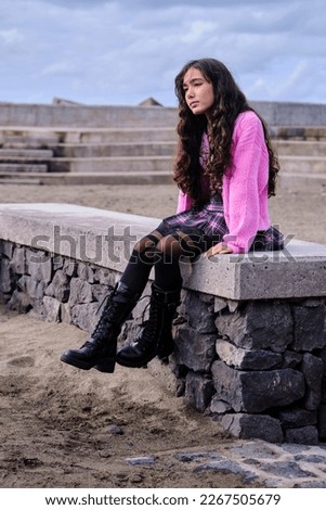 Profile portrait of teenage girl posing dressed in gothic style in black and pink colours sitting on a wall