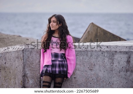 Gothic looking teenage girl posing next to wall with sea in background. She is wearing gothic clothes in black and pink colours with black boots.