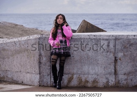 Gothic looking teenage girl posing next to wall with sea in background. She is wearing gothic clothes in black and pink colours with black boots.