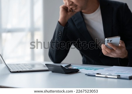 Young Asian businessman using mobile phone surfing the internet searching for financial information. Statistics, graphs, income charts. while working online on laptop computer at office.