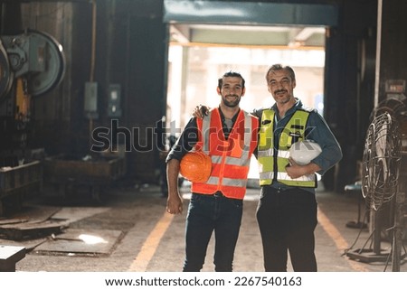 Portrait of senior and young male engineers and workers wearing safety vests and jacket while holding hardhat with arm around shoulder standing in front of machine in warehouse looking at camera Royalty-Free Stock Photo #2267540163