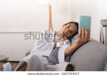 Young woman with the flu wearing a white robe, leaning on the couch, taking a picture for her social networks.