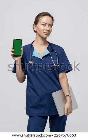 Portrait of a female surgeon in a blue suit on a white background. The doctor holds a laptop and a mobile phone with a green screen. The possibility of advertising