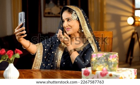 A happy Muslim lady in an ethnic dress posing clicking a selfie on her mobile - photogenic, a modern lifestyle . Colorful gift boxes are kept on the table on the occasion of the Eid festival 
