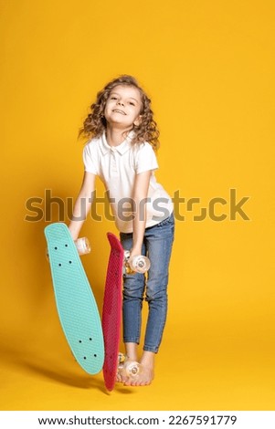 Full length of curly happy little girl in white polo, blue jeans who holding two colorful skateboards isolated on yellow background. Space for advertising 