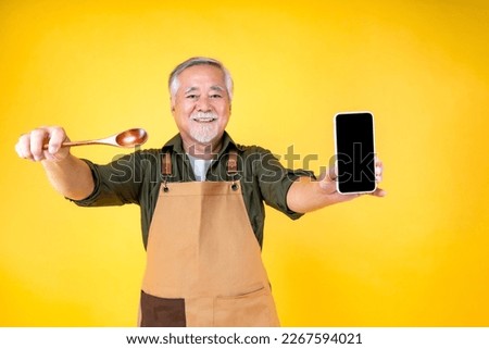 Portrait charming retired asian elder man has a white mustache and beard wearing apron holding fork spoon and smartphone order food in mobile online application isolated yellow background Royalty-Free Stock Photo #2267594021