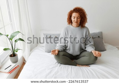 Pretty woman practicing yoga at home. Portrait of beautiful redhead caucasian plus-size female with curly hair sitting on bed in lotus posture, relaxing her mind, doing spiritual practice Royalty-Free Stock Photo #2267613537