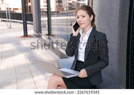 Business woman working on street. Young woman is sitting on bench with laptop and a mobile phone. Solving business problems against the backdrop of an office building in a business suit