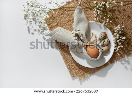 Minimalist Easter aesthetic table setting, egg decorated with bunny ears napkin on a white plate. Neutral floral background with copy space. Holiday branding spring template or banner design Royalty-Free Stock Photo #2267632889