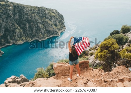Young woman standing on a rock cliff and waving the US flag while looking at sea beneath. Girl traveller waving the American flag while standing on a mountain top. 4 fourth of July Independence Day