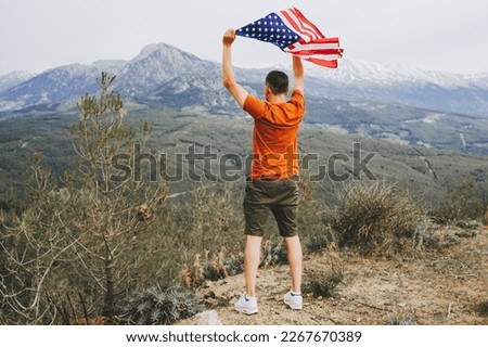 Young man standing on a rock cliff and waving the US flag while looking at mountains in the background. Male traveller waving American flag standing on mountain top. 4 fourth of July Independence Day.