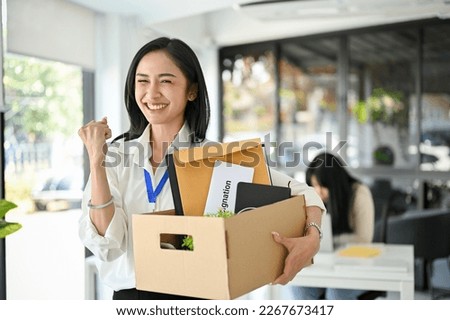Smiling and excited young Asian female office worker is celebrating her resignation, carrying her personal stuff, happy to leave her job or change job positions. Royalty-Free Stock Photo #2267673417