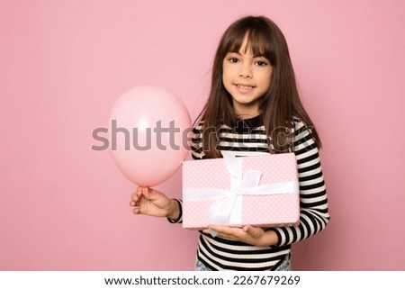 Picture of happy little girl child standing isolated over pink background. Looking camera holding gift box and balloon surprise.