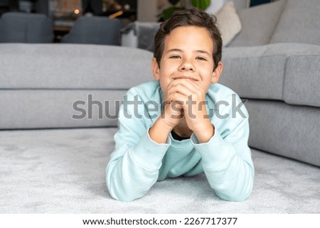 Happy childhood. Close up portrait of cheerful little boy smiling to camera, lying on floor at home, leaning chin on hands