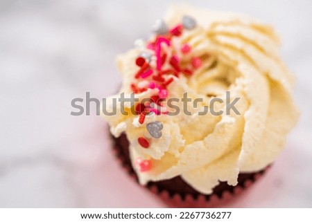 Piping white chocolate ganache frosting on top of red velvet cupcakes and topping with sprinkles.