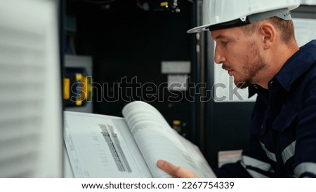 Caucasian production engineer in safety wear is reading the manual of a machine to find an error. A male factory worker is checking the industrial control panel of a robotic machine for maintenance. Royalty-Free Stock Photo #2267754339