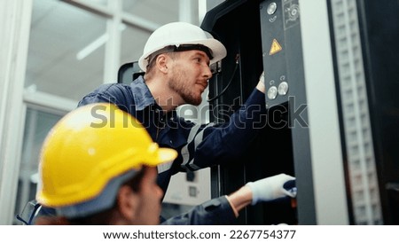 Two Caucasian production engineers in safety wear are assisting in adjusting and maintaining CNC machine in the factory. Male factory workers are examining the industrial machine to find an error.