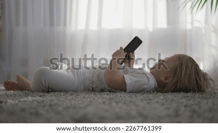 Cute infant looking into mobile phone screen lying down on carpet home
