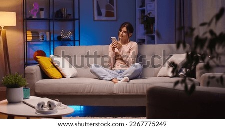Pretty Caucasian young woman in good mood typing and tapping on smartphone while resting on sofa at home. Beautiful happy female browsing on cellphone while sitting in room indoor. Leisure concept Royalty-Free Stock Photo #2267775249