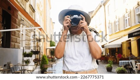Portrait of happy Asian beautiful woman in hat and glasses taking photo on photo camera while standing outdoor in town. Joyful pretty female tourist takes pictures in city. Traveler concept