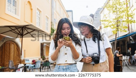 Portrait of cheerful Asian mother and daughter walking in town in good mood and looking around. Beautiful woman traveler in hat with photo camera. Young female tourist typing on smartphone in city