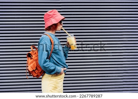 Stylish girl in bright clothes and bucket hat drinking sugar flavored tapioca bubble tea while walking near gray striped urban wall. Portrait of fashionable hipster girl. Street fashion. Copy space.