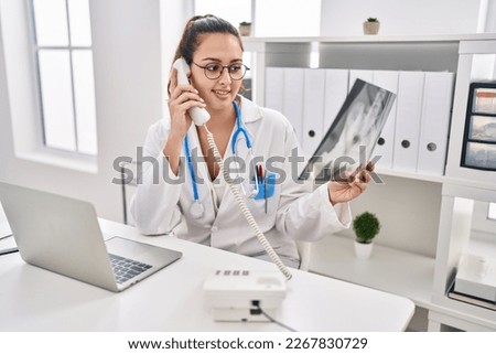 Young beautiful hispanic woman doctor talking on telephone holding xray at clinic
