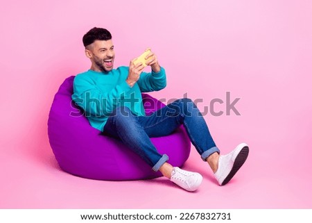 Full length photo of young nice guy sit bean bag playing mobile game excited dressed stylish blue look isolated on pink color background