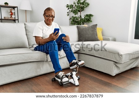 Senior african american woman using smartphone and doing sport using bike pedals machine at home Royalty-Free Stock Photo #2267837879