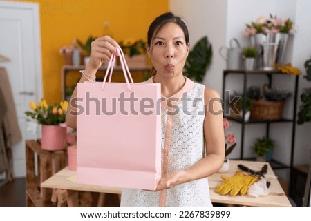 Middle age chinese woman working at florist shop holding shopping bag making fish face with mouth and squinting eyes, crazy and comical. 