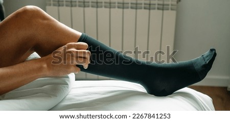a man puts on a black compression sock sitting on his bed at home, in a panoramic format to use as web banner or header Royalty-Free Stock Photo #2267841253