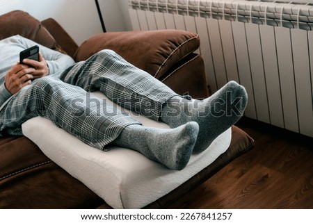 a man leans his legs on a leg elevation pillow, made of memory foam, while is resting on the sofa Royalty-Free Stock Photo #2267841257