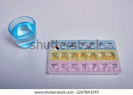 Top view of a plastic multi-colored convenient organizer for taking pills (vitamin) with the first open cell with medicines, next to it is a blue glass of water. Health care, reasonable medication.