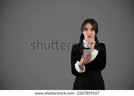 Pensive happy young girl student in black clothes with pigtails thinks to answer message in phone, looks up at empty space isolated on gray wall studio background. App for study, knowledge, learning