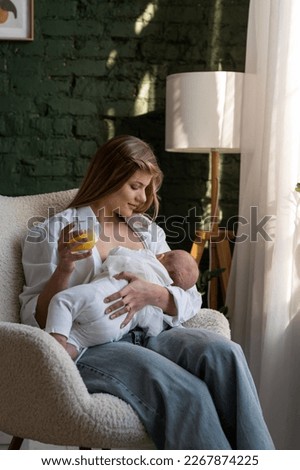 A young happy mother is feeding her baby while sitting in a soft chair in the living room and holding a glass of juice in her hand while looking at the newborn. Vertical photo.