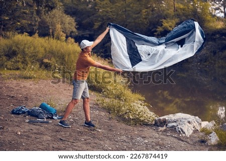 Man camping in nature, setting up the tent for overnight staying near forest river. Royalty-Free Stock Photo #2267874419