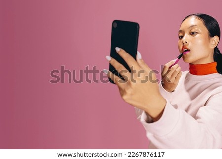 Fashionable female using a smartphone as a mirror to apply lipstick during her makeup routine. Young woman in her 20’s doing a bold two toned lip look, a stylish makeup trend.