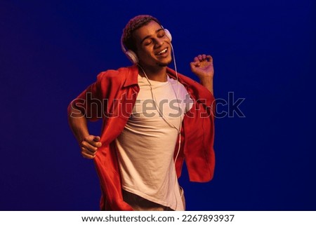 Excited smiling young african man in headphones dancing isolated over blue neon background
