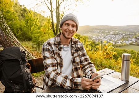 Young cheerful man tourist with backpack using mobile phone while sitting on wooden bench on hillside Royalty-Free Stock Photo #2267894137