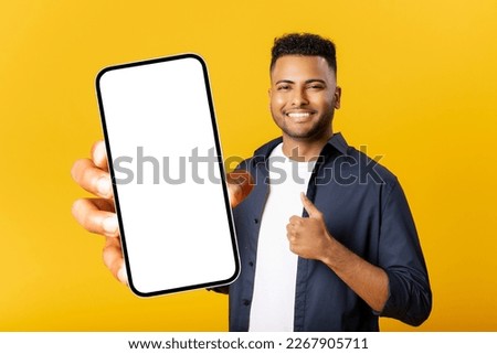 Cheerful Indian man with smartphone in the hand isolated on yellow background, showing smartphone with empty screen and thumbs up, advertising mobile app, showing deal
