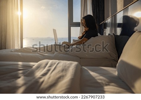 Happy fresh relaxed asian woman resting sitting on bed in hotel room with two beds using laptop enjoying good morning after wakeup in cozy room with soft light from window.  Royalty-Free Stock Photo #2267907233