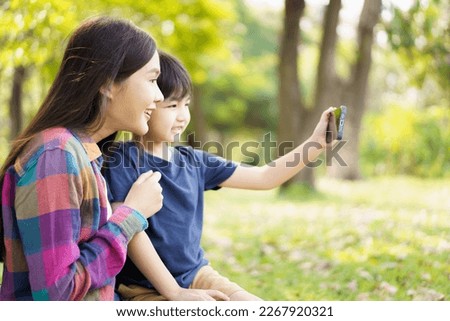 Happy cheerful Asian little boy using smartphone to take a selfie photography with his mother at the park in morning. Happy family time concept, woman staying with her little son in weekend.