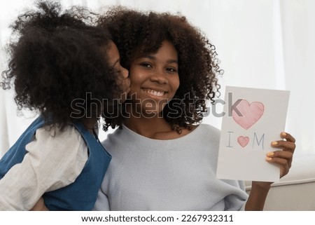 African American child girl hugging and kissing on mother cheek and holding mother day paper card at home