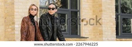 young couple in trendy sunglasses and leather jackets standing together outdoors, banner