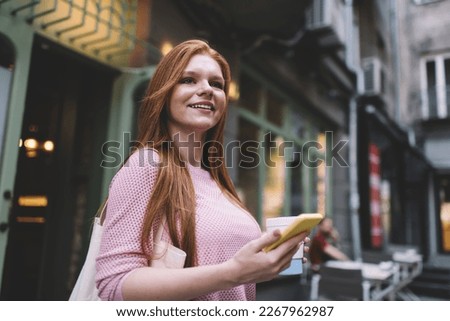 Side view of happy young female smiling and looking away with smartphone and coffee cup in hands while standing in daylight near cafe entrance and enjoying summertime