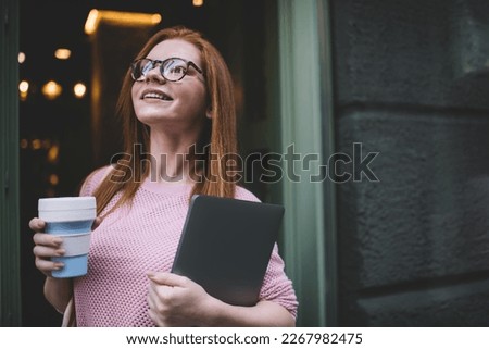 Happy adult female freelancer in casual outfit and eyeglasses standing near entrance while holding cup of takeaway coffee and tablet