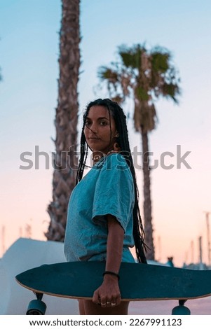 Pretty young woman with brown skin with long black braids posing with her skateboard dressed in a gray t-shirt and jean shorts strolling on the seafront in the summer evening in the evening light.