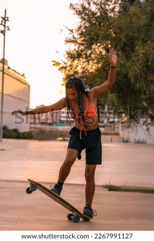 Pretty young woman with brown skin with long black braids skating with her skateboard dressed in an orange bikini and black shorts having fun on the seafront in the summer sunset in the warm light.